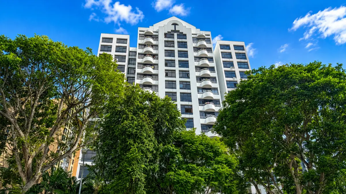 Modern white residential building with curved balconies rising behind dense green trees against a bright blue sky.