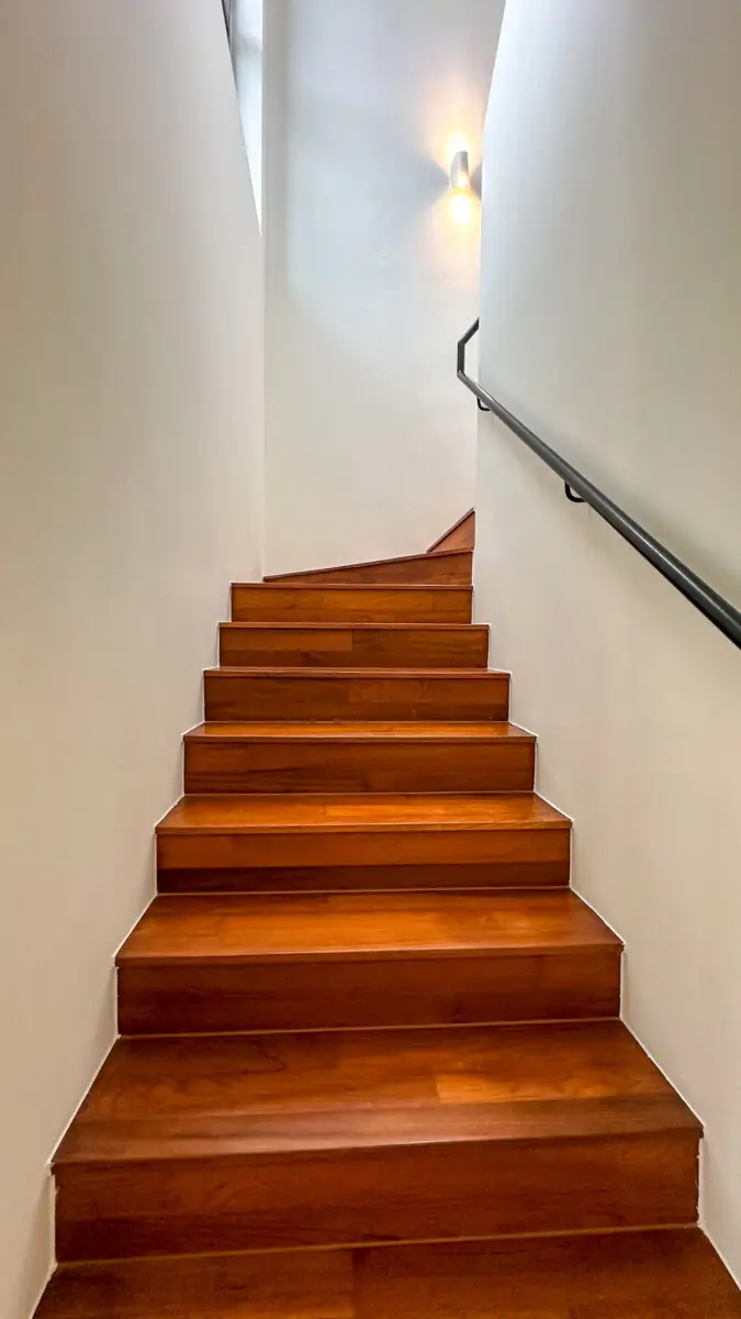 Narrow indoor wooden staircase ascending, with a black handrail on the right and a wall sconce near the top.