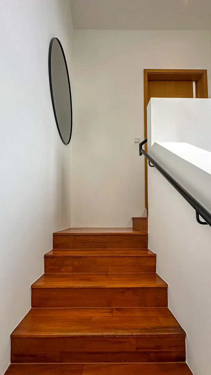 Wooden staircase against white walls with a round mirror on the left and a black handrail along the right, leading to a door at the top of the stairs.