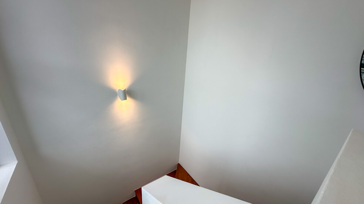 White interior corner with a wall sconce casting warm light on both walls near a wooden staircase.