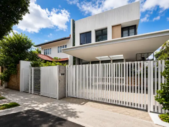 Modern two-story house with a tall white vertical-slat gate and a paved driveway under a bright blue sky in a sunny urban setting.