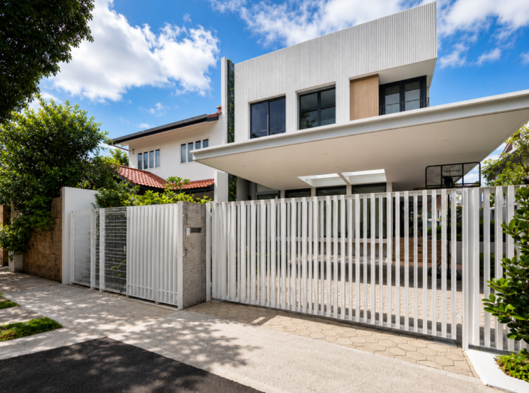 Modern two-story house with a tall white vertical-slat gate and a paved driveway under a bright blue sky in a sunny urban setting.