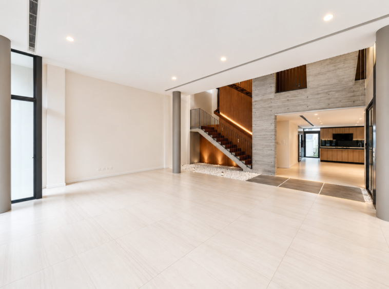 Spacious modern living area with large light tiles, a wooden staircase, and open-plan kitchen in the background.