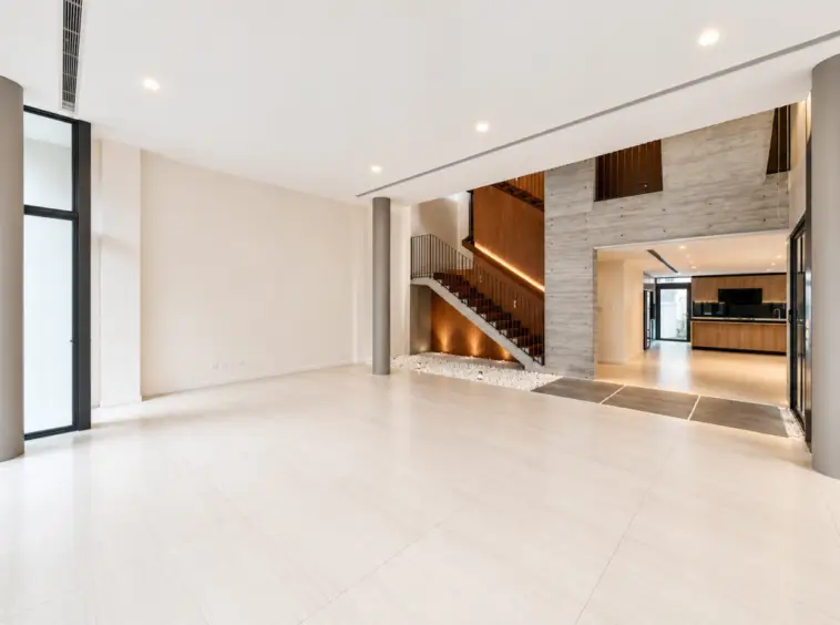 Spacious modern living area with large light tiles, a wooden staircase, and open-plan kitchen in the background.
