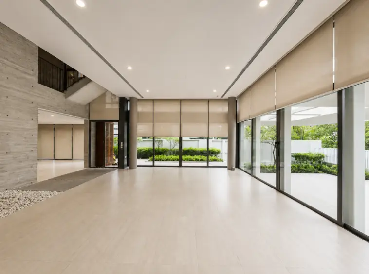 Bright modern lobby with wide glass doors, beige roller blinds, and light tiled floor; greenery visible outside.