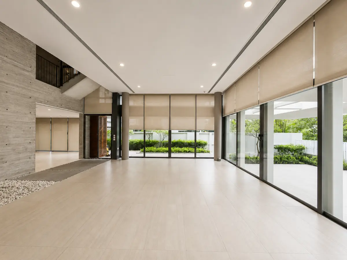 Bright modern lobby with wide glass doors, beige roller blinds, and light tiled floor; greenery visible outside.