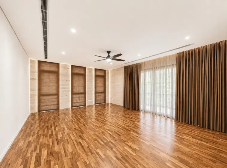 Spacious living room with hardwood floors, beige wall panels, and dark brown curtains over glass doors letting in light, plus a ceiling fan and recessed lighting.