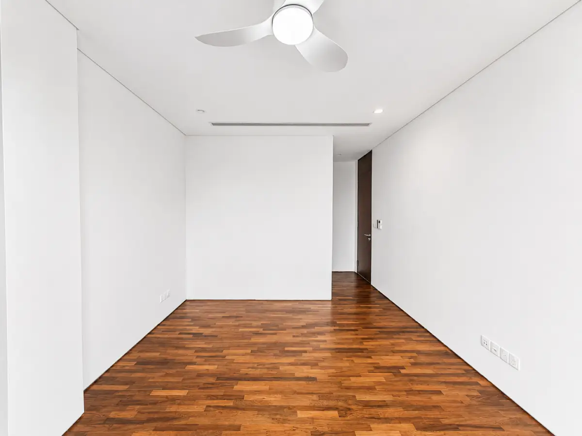 Empty white-walled room with a white ceiling fan, hardwood floor, and a dark wooden door at the far end.