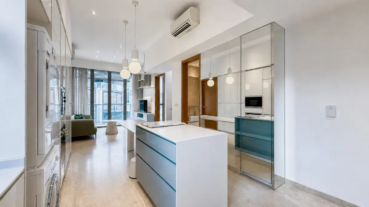 Open-plan kitchen with a white island featuring teal drawers, glass mirrored wall, and a sunny living area beyond.