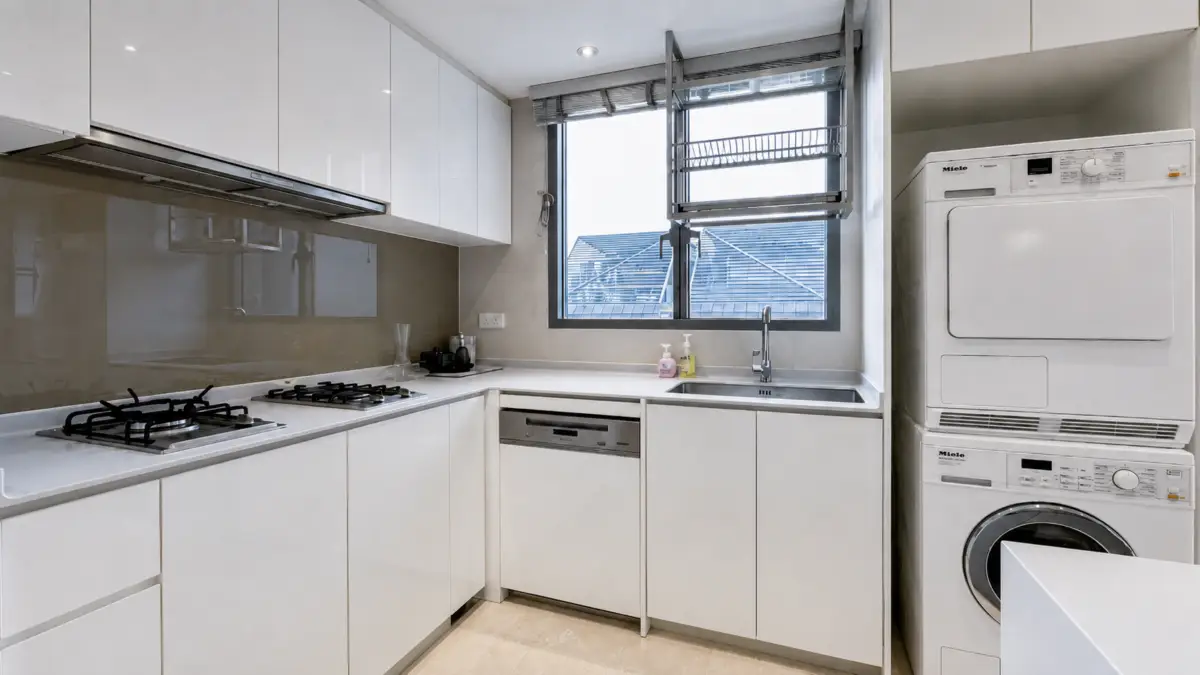 Modern white kitchen with a gas stove, built-in dishwasher, stainless sink under a window, and a stacked washer/dryer in the corner.