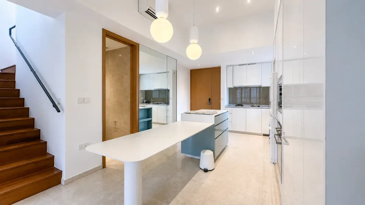 Open-plan kitchen with white cabinets, a central island with blue drawers, and wooden stairs on the left under pendant lights.