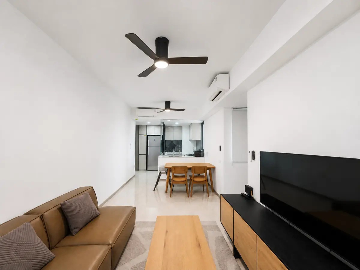 Bright, minimalist living area with a brown leather sofa, wooden coffee table, and a large TV on a black-and-wood console; dining table visible beyond. Uncluttered white walls and ceiling fans throughout.