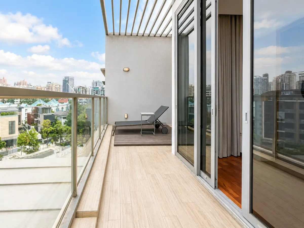 Apartment balcony with a gray chaise lounge and small side table, overlooking a city skyline through a glass railing under a blue sky.