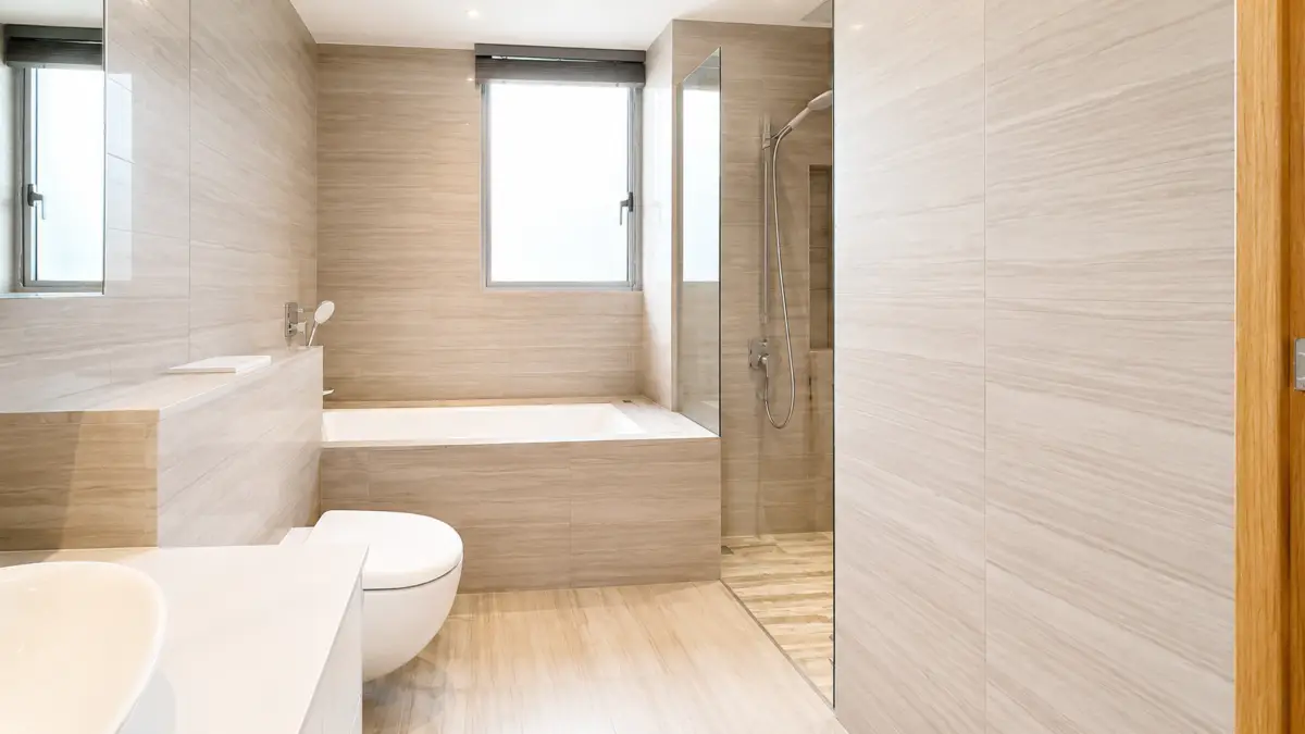 Bright beige tiled bathroom with built-in tub along back wall, glass shower to the right, and a white toilet in the foreground.
