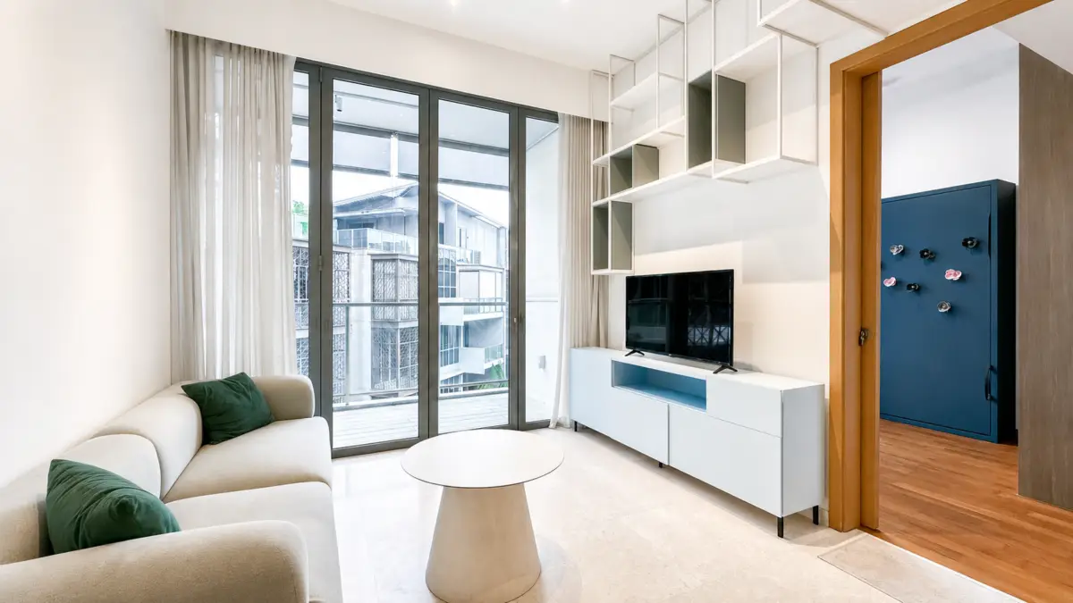 Bright modern living room with a beige sofa and green cushions, round white coffee table, and a TV on a white media unit by large glass doors to a balcony.