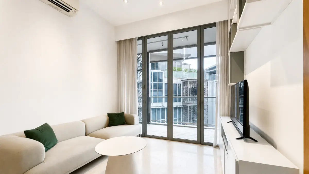 Bright, modern living room with a curved cream sofa, two dark-green cushions, and a round white coffee table facing a TV on a white media console beneath large glass balcony doors.