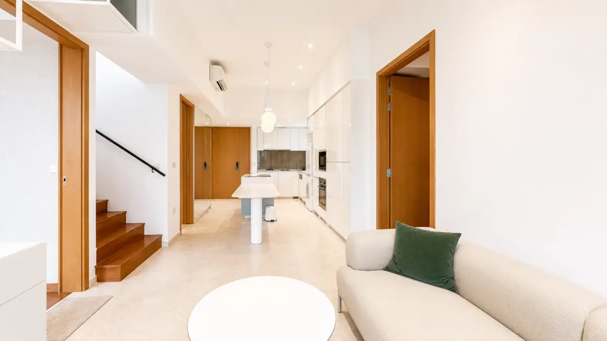 Modern open-concept living space with white kitchen, island, and beige sofa with green pillow; wooden doors and stairs on the left