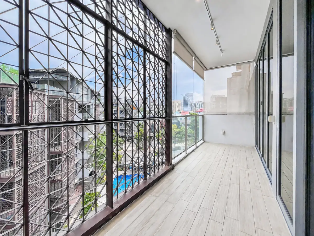 Balcony with light wood-like flooring, glass railing, and a metal lattice screen on the left; city buildings visible in the distance.