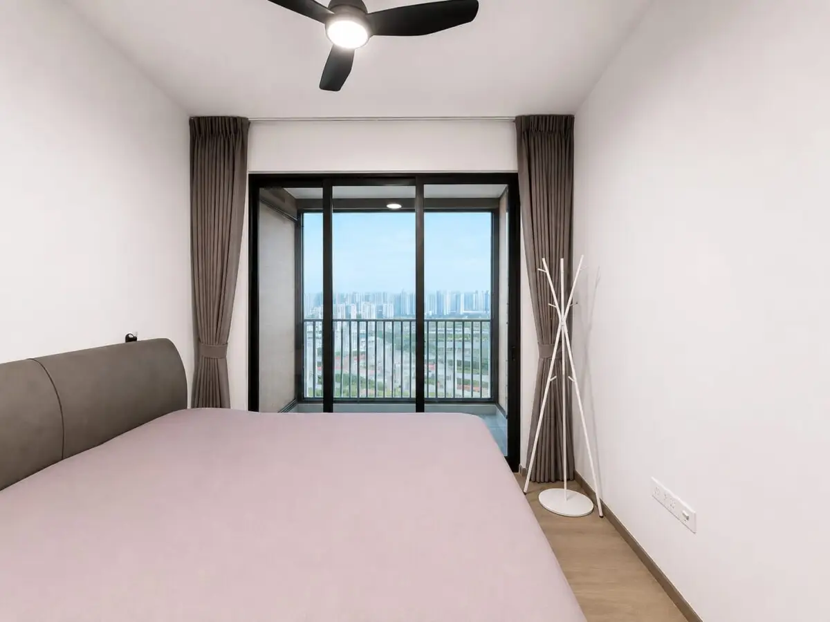 Bedroom with a pink duvet, brown headboard, and white walls; glass balcony doors open to a city skyline.