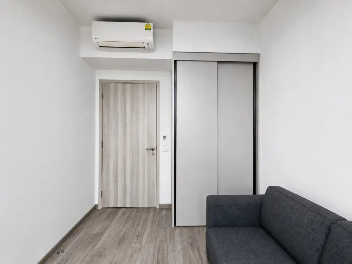 Living room with a light wood door, sliding closet doors, wall-mounted air conditioner, and a gray sofa in a minimalist space