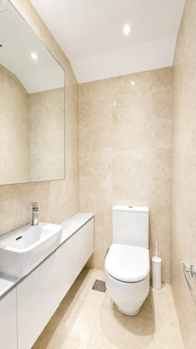 Modern beige-tiled bathroom with wall-mounted white vanity, rectangular sink, large mirror, and a white toilet nearby.