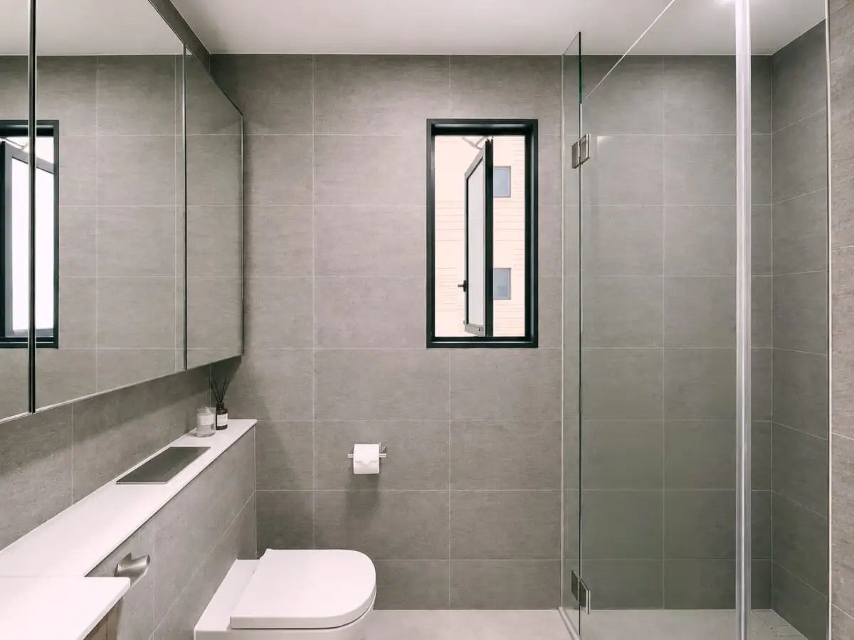 Minimalist gray-tiled bathroom with a wall mirror, open window, wall-mounted toilet, and glass shower enclosure.