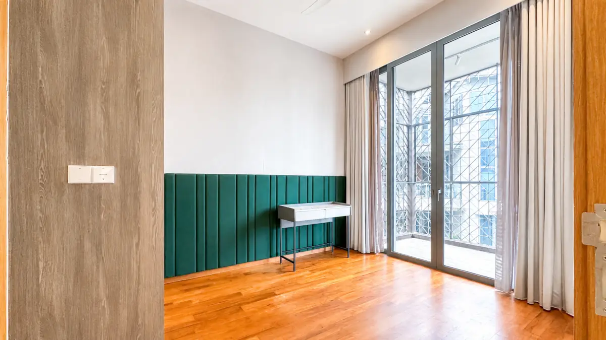 Modern minimalist room with teal vertical paneling along the wall, a small white desk, and a large glass balcony door with curtains; warm wood floor.