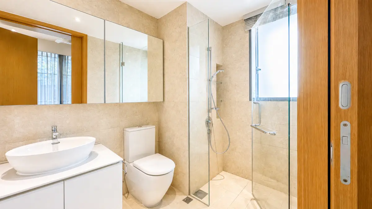 Modern beige tiled bathroom with a glass shower enclosure, white vessel sink, and toilet beneath mirrored cabinets.