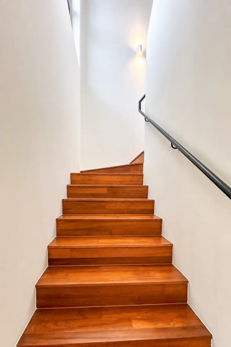 Wooden stairs rise between white walls with a dark handrail along the right side.] ,...