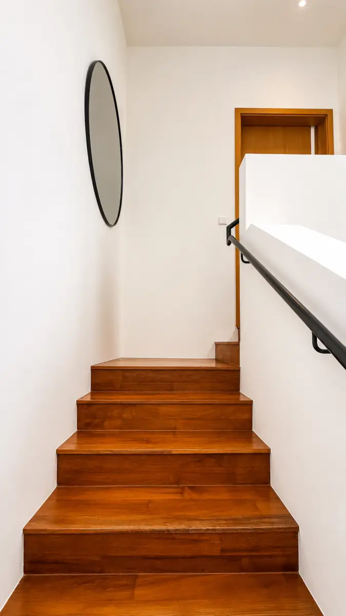 Wooden staircase with white walls, a black handrail on the right, and a round mirror on the left wall in a bright interior.