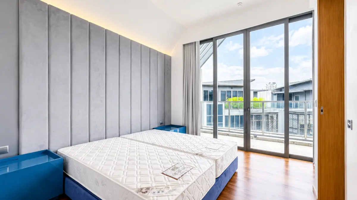 Contemporary hotel bedroom with two mattresses on a blue platform, a tall grey padded headboard, and glass balcony doors-out to a city view.