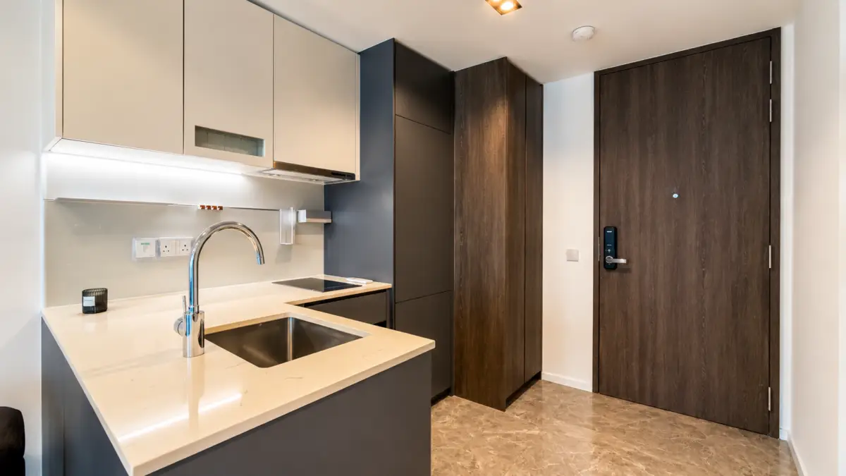 Modern kitchenette in an apartment hallway with a marble countertop, sink, and built-in appliances beside a dark wood storage cabinet and front door.