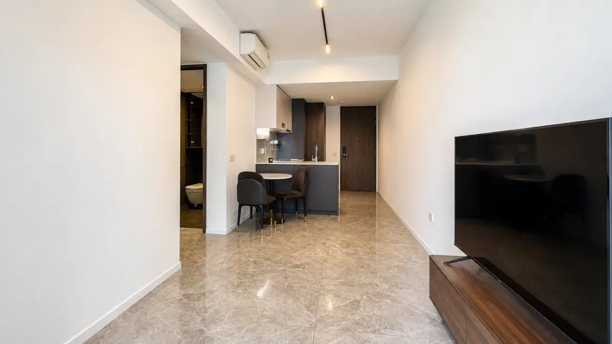 Open-plan living area with kitchenette, round dining table and three chairs, marble floor, and a large TV on a wooden stand.