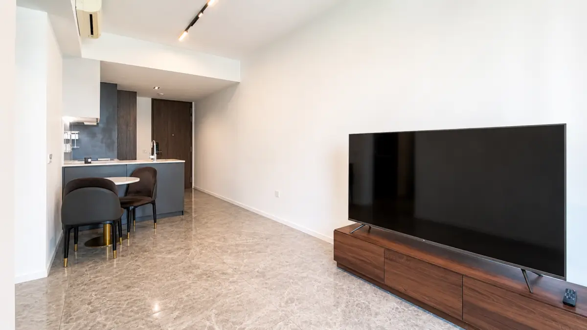 Open-plan living area with a blue-gray kitchen, a small round table and two chairs, and a large TV on a wooden stand.