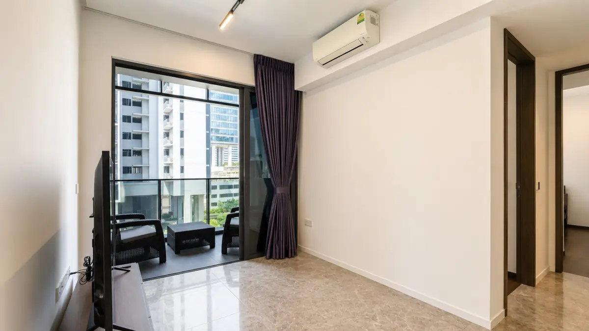 Living room with a flat-screen TV on a stand, marble floor, and glass sliding door opening to a balcony with two black wicker chairs and a small table; purple blackout curtains and an overhead air conditioner.
