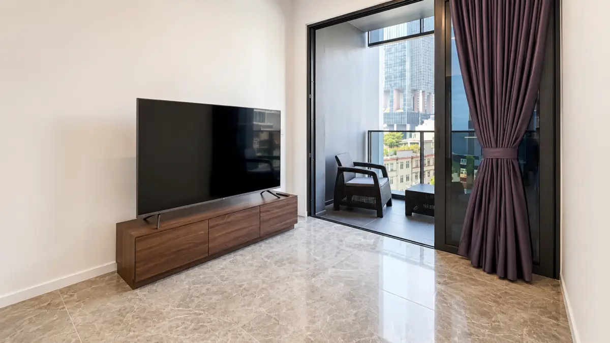 Living room with a large flat-screen TV on a wooden console and a marble floor, opening to a balcony with dark purple curtains.