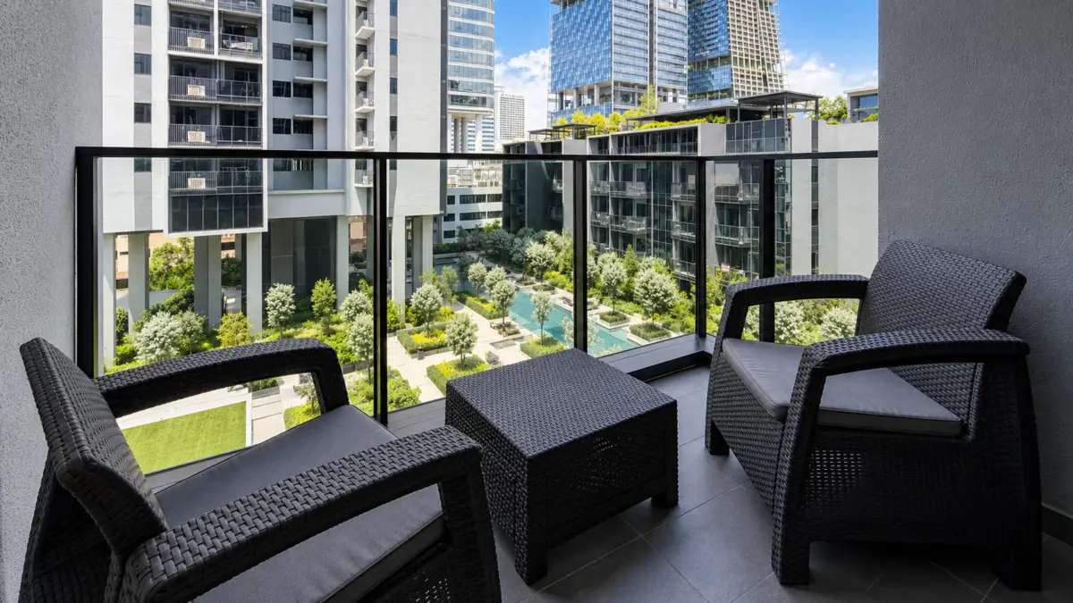 Balcony seating area with two dark wicker chairs and a table, overlooking a landscaped courtyard between modern high-rise buildings.