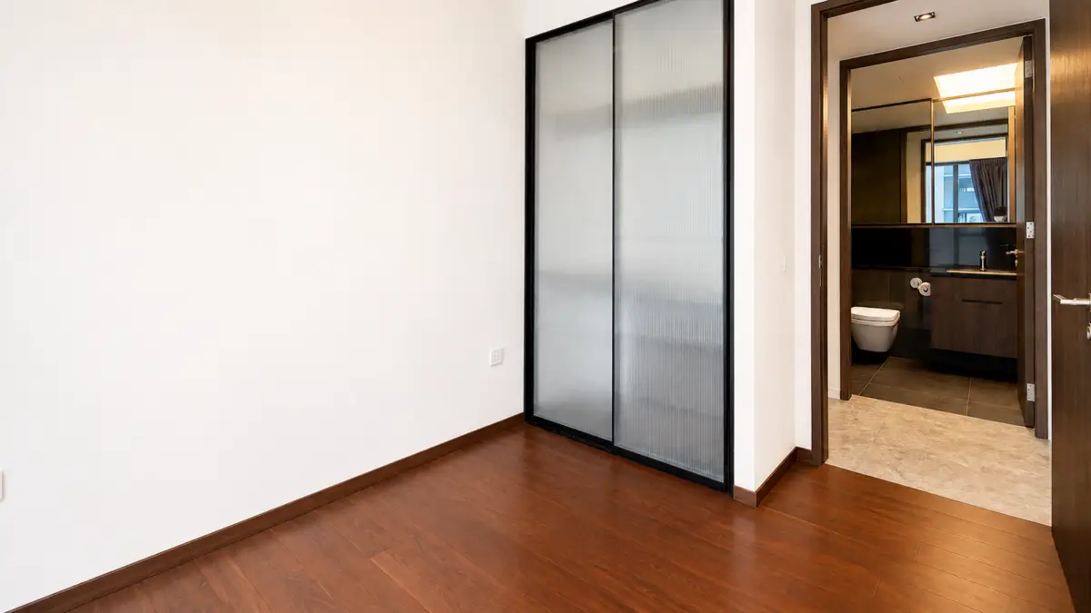 Empty room with dark wood floor, frosted glass sliding closet, and an open doorway to a bathroom with a toilet and sink.